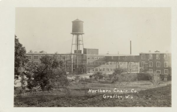 View toward the factory with a water tower and an elevated walkway connecting the buildings. Caption reads: "Northern Chair Co., Grafton, Wis."