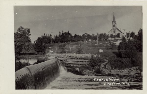 View of Grafton from across a dam on the Milwaukee River. St. Joseph's Catholic Church is in the distance on a hill. Caption reads: "Scenic View, Grafton, Wis."