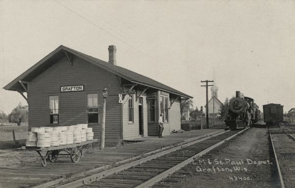 View across railroad tracks towards the Grafton Depot and an oncoming train. A man is standing on the platform. St. Paul's Lutheran Church is in the background.
