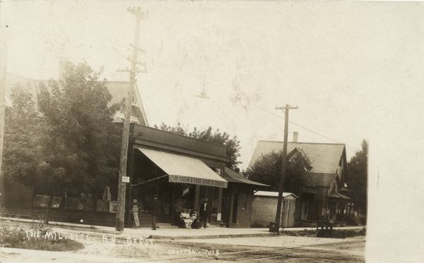 Text on front of postcard reads: "The Milw. Elec. Ry. Depot". View towards a railroad crossing and an ice cream, drug and candy store on the corner with show windows and a striped awning. Children and a woman are sitting on a bench in front, and a man is standing next to them.