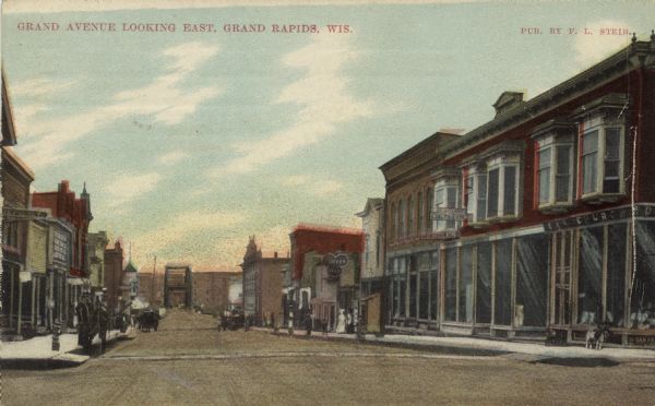 Hand-colored view of Grand Avenue looking east toward the bridge. The street is lined with businesses. Caption reads: "Grand Avenue Looking East, Grand Rapids, Wis."