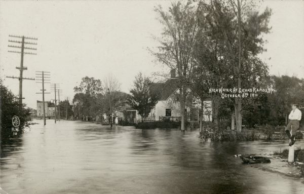 View of a flooded street in a residential neighborhood. A boy is sitting on a post in the foreground on the far right. Caption reads: "High Water at Grand Rapids, Wis. October 8th, 1911."