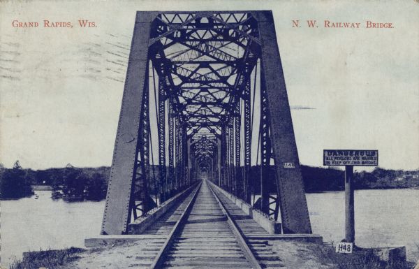 View down the tracks through the railroad bridge over the Wisconsin River. There is a warning sign on the right at the entrance. Caption reads: "Grand Rapids, Wis. N.W. Railway Bridge."