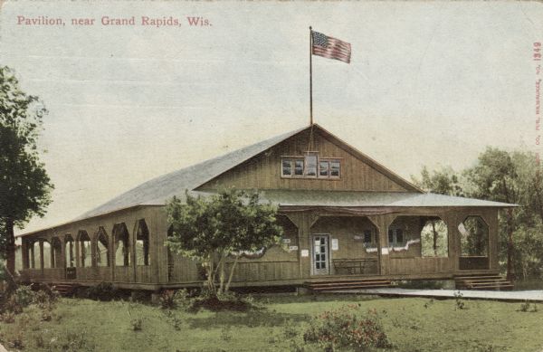 Hand-colored postcard of an enclosed pavilion with a wrap-around porch. A flag is flying from a flagpole on the roof. Caption reads: "Pavilion, near Grand Rapids, Wis."