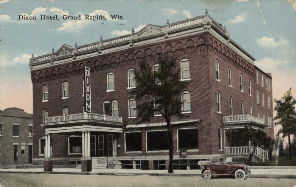 Hand-colored postcard of the Dixon Hotel. A large sign is above the entrance, and a bar is in the basement. A car is parked at the curb on the right. Caption reads: "Dixon Hotel, Grand Rapids, Wis."