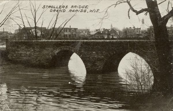 View from shoreline towards a stone pedestrian bridge on a river. The city skyline is in the distance. Caption reads: "Strollers Bridge, Grand Rapids, Wis."