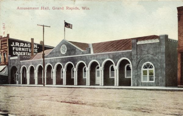 View across street towards an arcade. A furniture store/undertaker is next door. Caption reads: "Amusement Hall, Grand Rapids, Wis."