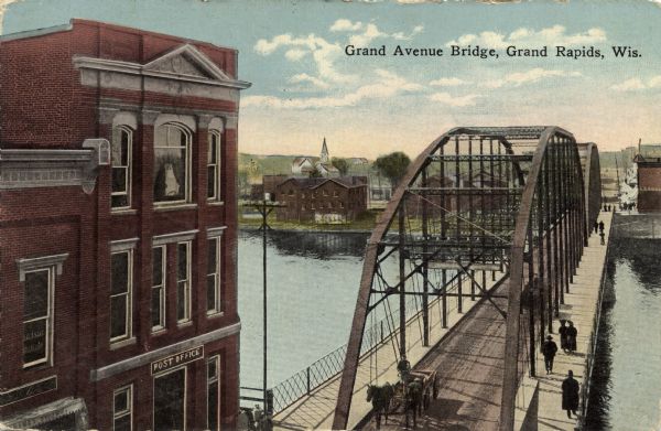 Elevated view of the Grand Avenue Bridge. A horse and wagon is coming across the bridge, and pedestrians are crossing on the walkway. There is a post office in the left foreground. Caption reads: "Grand Avenue Bridge, Grand Rapids, Wis."