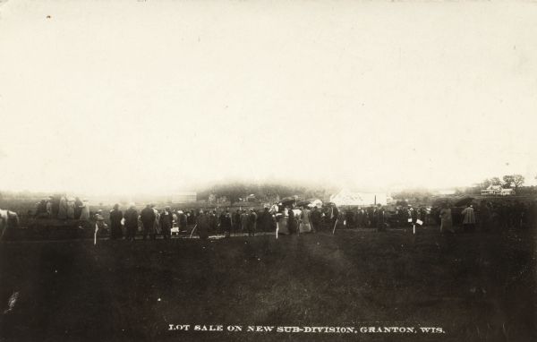 View toward a group o people gathered at a vacant lot. There are dwellings in the distance. Caption reads: "Lot Sale on Sub-Division, Granton, Wis."
