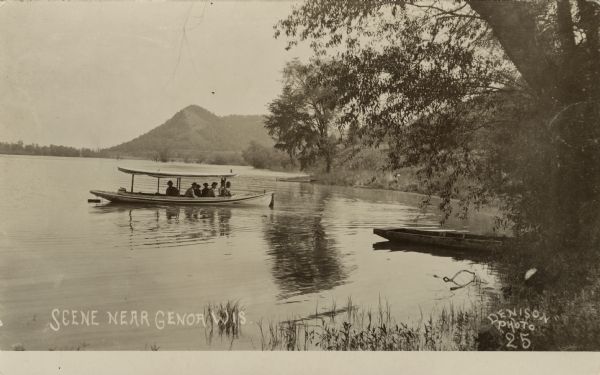 View from shoreline towards a small group of people on an excursion boat in the water near the shore. Bluffs are in the background. Caption reads: "Scene near Genoa, Wis."