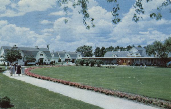 Kodachrome postcard of the grounds and buildings of the Honey Bear Farm. A group of women walking along a pathway lined with flowers. Text on reverse reads: "On the shores of Powers Lake".