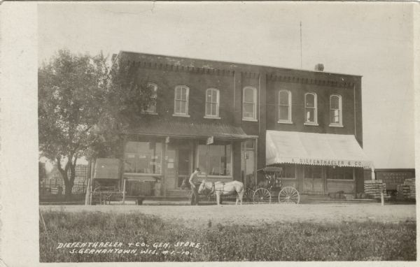 View of A. Diefenthaler's General Store and the building next to it. A man is standing near a horse and buggy parked in the street. Freight cars are behind the building. Caption reads: "Diefenthaler & Co. General Store, S. Germantown, Wis."