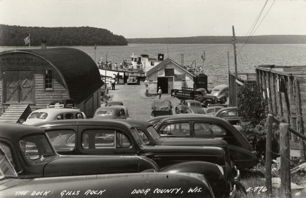 View of the excursion boat dock from the parking lot. Several automobiles and trucks are parked in the foreground. An excursion boat with tourists on board is docked at the pier. Caption reads: "The Dock, Gills Rock, Door County, Wis."