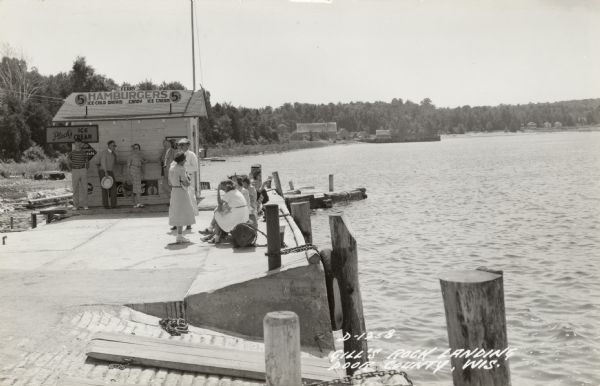 View down the Gills Rock boat landing with tourists gathered waiting for a ferry. A hamburger stand is at the end of the dock. Caption reads: "Gills Rock Landing, Door County, Wis."