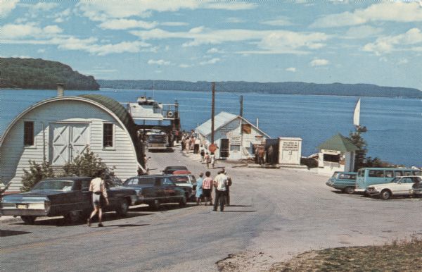 View down road towards the ferry dock at Gills Rock. A ferry is unloading cars. A line of cars and a group of people are waiting to get on the ferry.