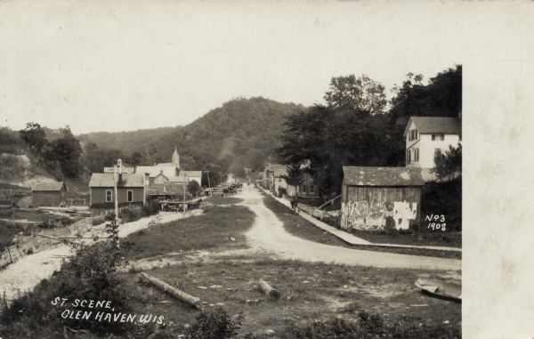 View of a street with a lumber office and a church on the left, and dwellings on the right. A man and a dog are on the sidewalk. A tree-covered hill is in the distance. The remains of a Ringling Brothers poster is on the side of a building on the right. Caption reads: "Street Scene, Glen Haven, Wis."