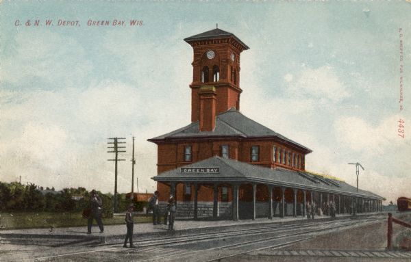 View of the train depot with a clock tower. Men are gathered on the platform.