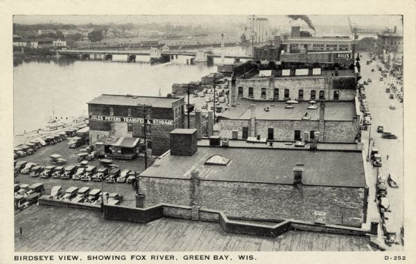 Elevated view of the Fox River at Green Bay. Warehouses and factories are along the riverfront, and a drawbridge is spanning the river in the background.
