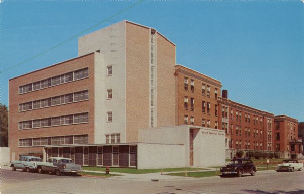 Color reproduction of a four-story brick hospital with cars parked at the curbs. A fire hydrant is on the corner.
