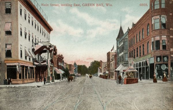 Hand-colored view of the businesses along Main Street. Streetcar tracks run down the street, and pedestrians are on the sidewalks. Caption reads: "Main Street, Looking East, Green Bay, Wis."