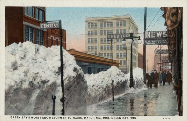 View of a city sidewalk after a snow storm, with tall piles of snow along the curb. Men are walking along the sidewalk. Caption reads: "Green Bay's Worst Snow Storm in 40 Years, March 9, 1918, Green Bay, Wis."