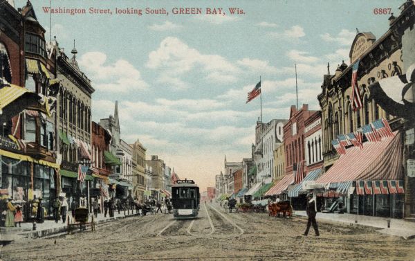 Hand-colored view of a city street lined with buildings. A streetcar and horse-drawn vehicles are in the street. People are on the sidewalks, and many flags are flying from buildings. Caption reads: "Washington Street, Looking South, Green Bay, Wis."