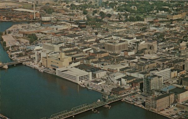 Color postcard of downtown Green Bay and bridges spanning the Fox River. The Walnut Street Bridge is in the foreground. A 4-level parking garage is on the waterfront.