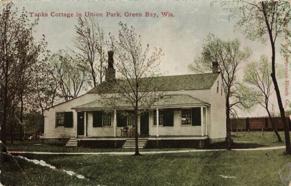 View across lawn towards the front of the cottage. Train cars are in the background. On the original site on the west side of the Fox River, before restoration. Caption reads: "Tanks Cottage in Union Park, Green Bay, Wis."
