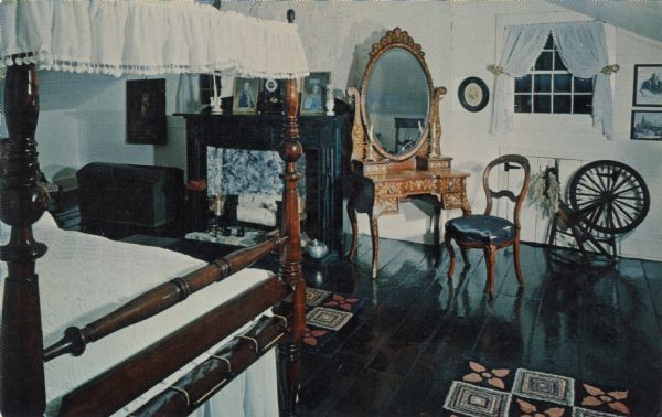 Interior color postcard of a bedroom at the Tank House built in 1776. In the room is a bed, chest, fireplace, dressing table with mirror and a spinning wheel.