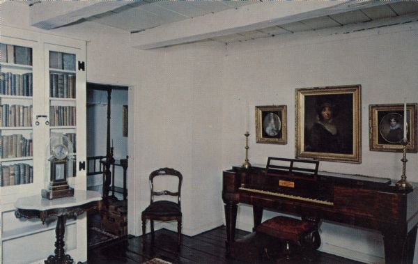 Interior color postcard of the library in the Tank House, built by Joseph Le Roi in 1776. A piano is against the wall on the right under framed portraits. On the left is a clock on a table in front of a books in a glass door fronted built-in cabinet near an open doorway.