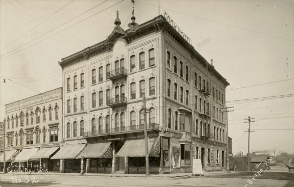 View across intersection towards the Cook Hotel. There are rod iron balconies above the entrance, and fire escapes are on the side. Caption reads: "Cook Hotel, Green Bay, Wis."