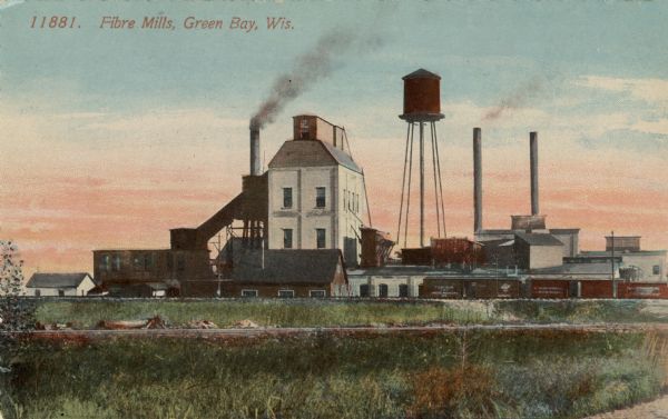 View of the mill just beyond railroad tracks, with smokestacks and a water tower. Caption reads: "Fibre Mills, Green Bay, Wis."