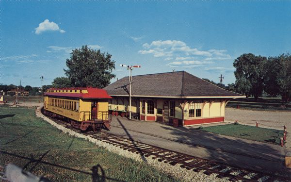 Color postcard of a yellow train car next to a yellow depot.

Text on reverse reads: "National Railroad Museum Green Bay, Wisconsin
Internationally famous as a shrine to preserve the story of American railroading. Both standard and narrow gauge steam trains are in operation. Children of all ages will delight in riding the steam trains."