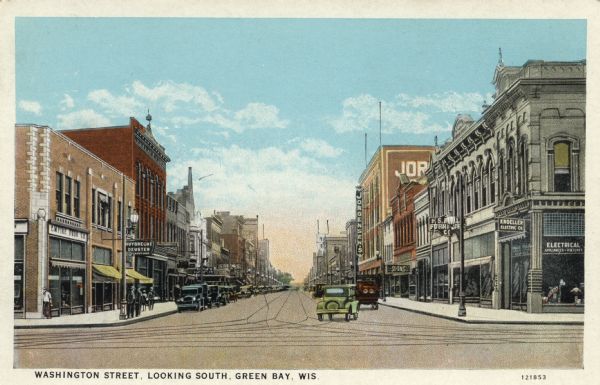 Colorized postcard view of Washington Street at an intersection. Commercial buildings are on both sides of the street, with automobiles parked along the curbs, and streetcar tracks run down the middle of the street. Caption reads: "Washington Street, Looking South, Green Bay, Wis."