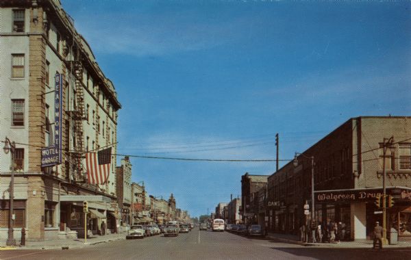 Colorized postcard view down a street in a main shopping district. Traffic signals are on the corners, and a large flag is hanging from the Beaumont Hotel on the left. A Walgreen Drug store is on the corner onthe right.