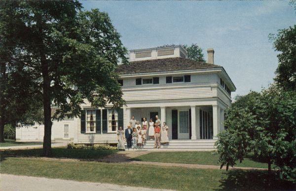 Color postcard view towards the Butternut House. A tour guide and touring group are standing on the front steps of the porch.

Text on reverse reads: "The Butternut House, so called because of the extensive use of native butternut for both exterior and interior work, was the home of Charles and Julia (Wade) Robinson. Robinson operated the sawmill and is believed to be responsible for the architectural design of Wade House as well as that of his own home and the Methodist Church across the street."