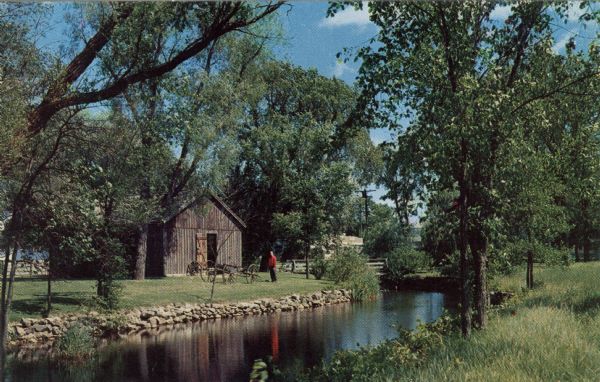 Ektachrome postcard of the blacksmith shop next to a stream.

Text on reverse reads: "The Blacksmith Shop was Sylvanus Wade's first venture in Greenbush. As stagecoach traffic increased he decided to build an inn also. Up the river from the smithy can be seen the remains of an old dam. On the left bank, looking upstream, stood the sawmill, on the right bank, a gristmill."