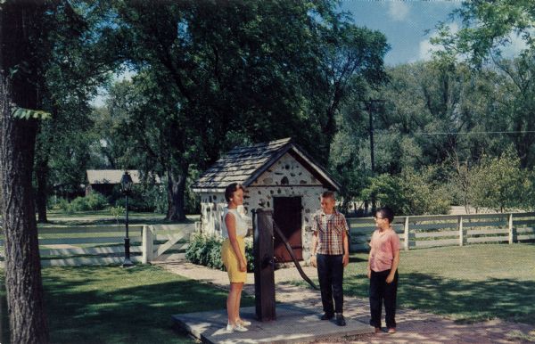 Ektachrome postcard of a group of people standing near the smokehouse and water pump.

Text on reverse reads: "Outside the summer kitchen stands the old pump and from the smokehouse just beyond came the hams and bacon to provide a breakfast for which Wade House was famous. From a gristmill on the nearby Mullett River came buckwheat flour and the Sugaring Cabin the maple syrup."