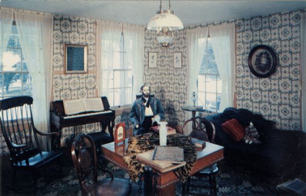Ektachrome postcard of the Green Parlor, with a mannequin of a man in period dress sitting in a chair.

Text on reverse reads: "The Green Parlor or sitting room is on the second floor. As the stagecoach traffic declined the upstairs rooms sometimes were rented as apartment units. The melodeon was purchased at Fond du Lac for $60 in 1864 for Allie Stannard. Allie, then employed at the inn at $12 a month 'and keep' later married Hollis Wade."