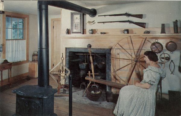 Ektachrome postcard of the kitchen. A woman wearing period clothing is sitting by a spinning wheel near the woodstove and fireplace.

Text on reverse reads: "The Kitchen contains the only fireplace in Wade House. Sylvanus Wade wanted to 'go modern' and install stoves throughout, but Betsy insisted on a fireplace in her kitchen. Here the food was prepared for the hungry travelers and the Wades' own large family. Over the mantle are the guns used to bring home game for the inn's table."