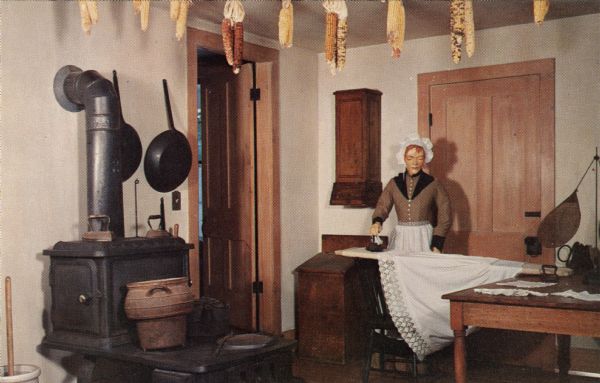 Ektachrome postcard of a mannequin of a women wearing period dress ironing in the summer kitchen. Dried corn is strung up near the ceiling.

Text on reverse reads: "The Summer Kitchen has a wood burning stove of unusual design. Many visitors find the collection of stoves through the inn highly interesting. The mannequin is busy ironing and her finished work on the clothes rack includes the wedding nightgown of Allie Wade."