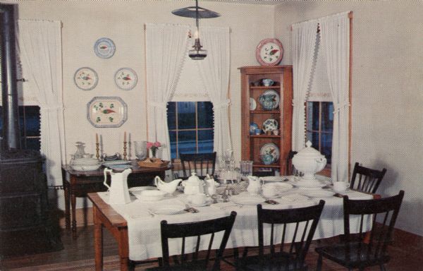 Ektachrome postcard of the dining room with the table set with dinnerware.

Text on reverse reads: "The Dining Room tables are set with the original ironstone dinnerware. There is also a fine collection of rare spatterware of the pea fowl pattern made in Staffordshire, England. The table and chairs, too, are part of the original furnishings of the old inn, which back in the stagecoach days was considered 'the best on the road.'"
