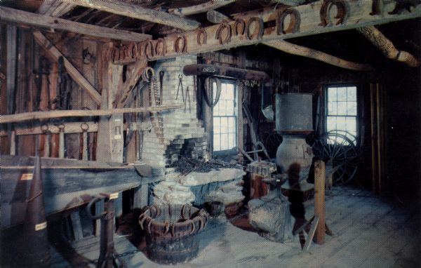 Ektachrome postcard of the interior of the blacksmith shop.

Text on reverse reads: "In the blacksmith shop the giant bellows and forge are in fully operable condition. The shop also contains an 'ox sling' by which oxen were lifted to be shod because they were unable to stand on three legs."