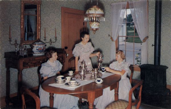 Three women in period clothes are having tea in the dining room.

Text on reverse reads: "Dining Room, Butternut House, Old Wade House State Park, Greenbush, Wis. In contrast to the simple furnishings of the Wade House, Butternut House, also dating to the 1850s, contains the more luxurious furnishings of the village mansion of that era."