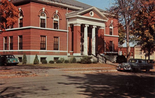 Ektachrome postcard of the facade of the Green Lake County Courthouse. An automobile is parked at the curb, and another automobile is in the side driveway.