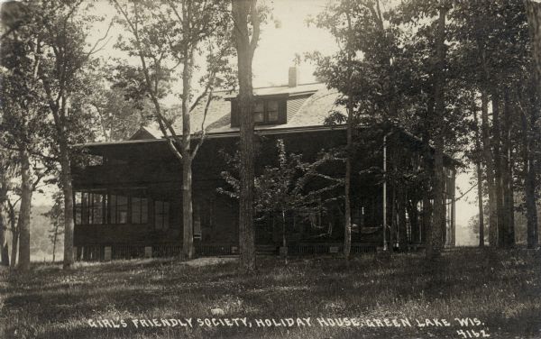 View across lawn towards a two-story lodging. The building has screened porches on the first and second floors. Caption reads: "Girl's Friendly Society, Holiday House, Green Lake, Wis."