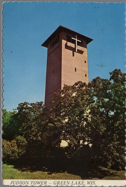 View across lawn and through trees towards a standing tower, with small windows and a cross at the top under the roof overhang. Caption reads: "Judson Tower, Green Lake, Wis."

Text on reverse reads: "Judson Tower overlooks the lake and provides an impressive view of the entire Assembly grounds."