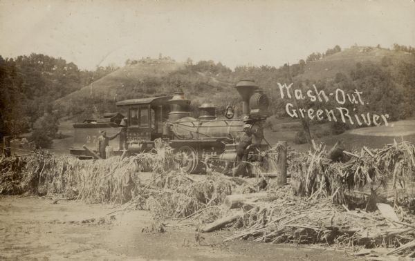 View of a locomotive stopped in a wash-out. Two men are standing nearby. Bluffs are in the background. Caption reads: "Wash-Out, Green River, Wis."