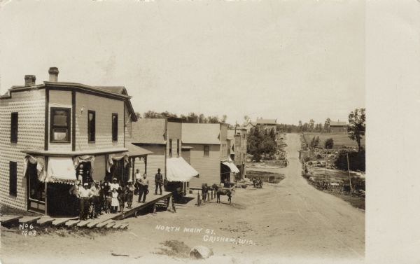 Elevated view of a street scene, with businesses on the left. A group of people are standing on the porch of the corner store. A man is standing near a horse tied up at a rail in front of one of the storefronts further down the street. Horse-drawn vehicles are parked on the right side of the street. Caption reads: "North Main Street, Grisham [sic] , Wis."