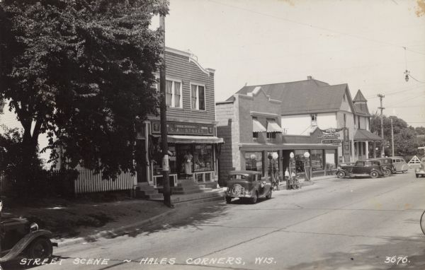 View of a central business district, with an I.G.A. Grocery Store, a filling station, a Dodge car dealer, and a tavern. Automobiles are parked at the curb. Two boys and a bicycle are near the gas pumps. Caption reads: "Street Scene ~ Hales Corners, Wis."